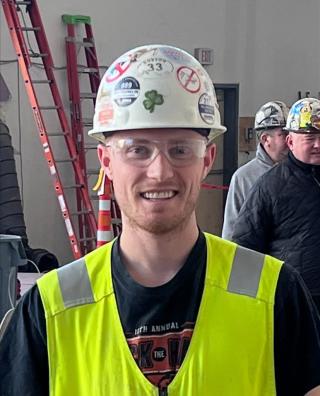 Sean Arnstein on a construction site, wearing a white hard hat covered with stickers, safety glasses, and a bright yellow safety vest.