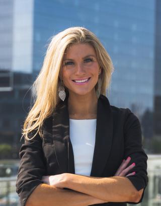 Portrait of Lindsay Higgins on a city boardwalk, wearing a black blazer over a white dress, with glass office towers and a marina in the background.